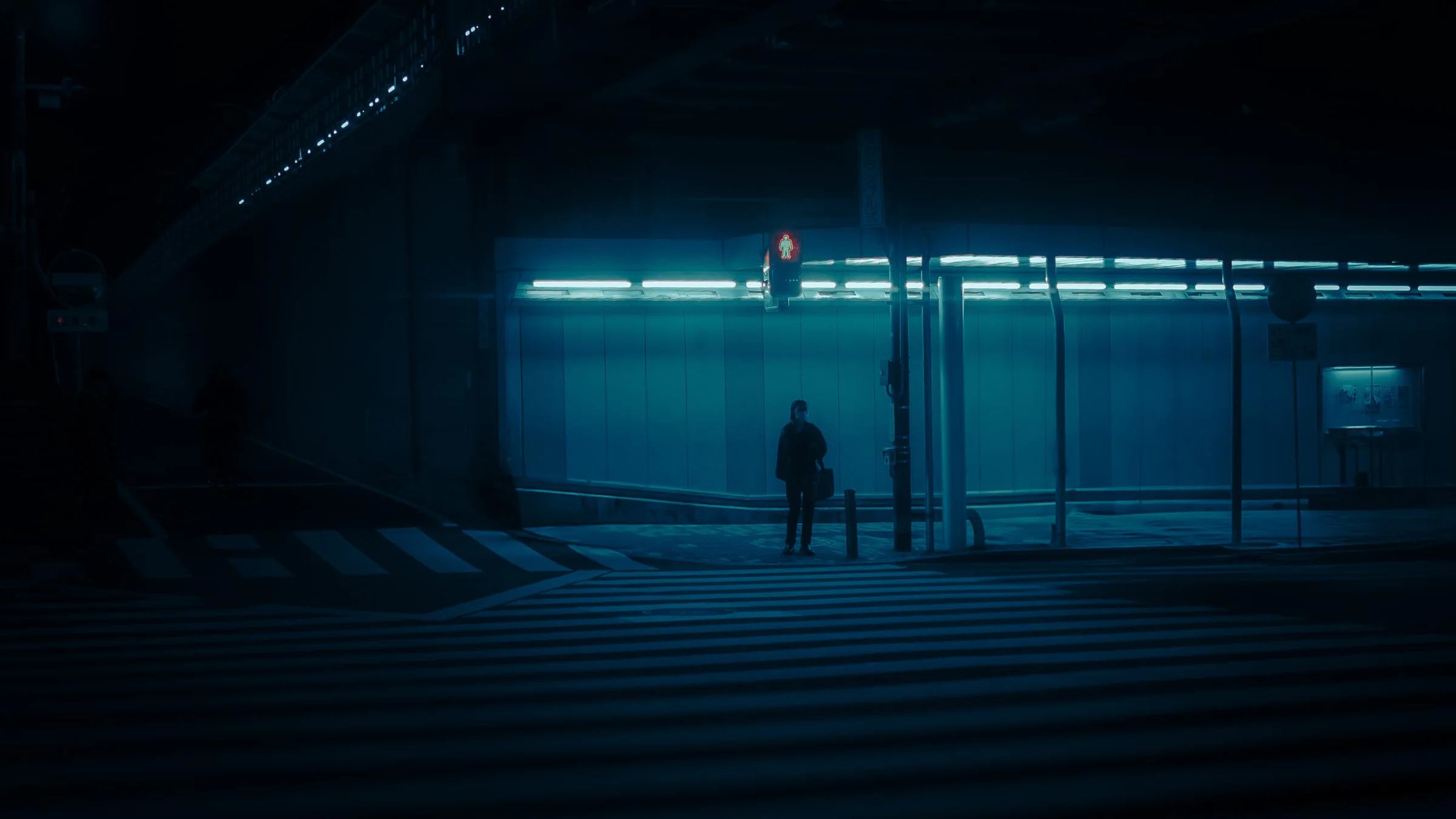 A lone figure waits under a red pedestrian light in front of a wall lit by blue neon, at night in Tokyo